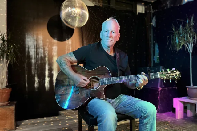 Lars Bo Bendixen sidder med en akustisk guitar på en lille scene med discokugle og planter og kigger ind i kameraet