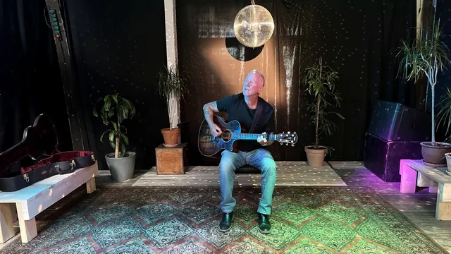 Lars Bo Bendixen spiller guitar på en lille scene med tæpper, planter og en guitarkasse