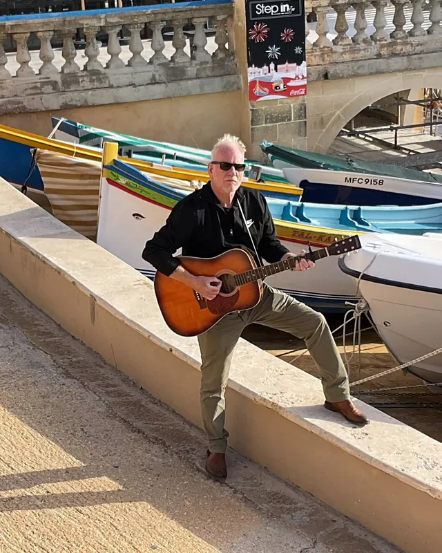 Lars Bo Bendixen med akustisk guitar foran fiskerbåde på Malta