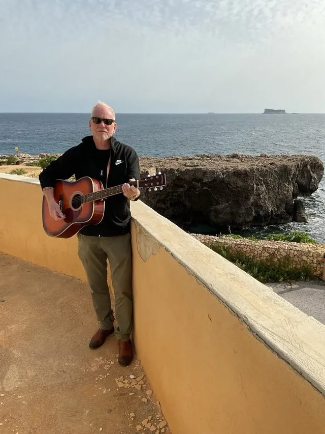 Lars Bo Bendixen med akustisk guitar lænet mod en gul mur med havet bagved